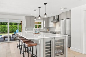 A modern kitchen with a large island, bar stools, stainless steel appliances, and a sliding glass door leading to an outdoor area.