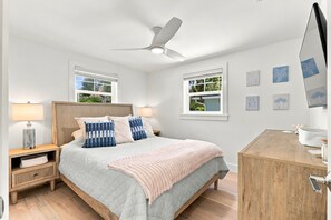 A bedroom with a bed, bedside tables, a desk, a ceiling fan, and windows.