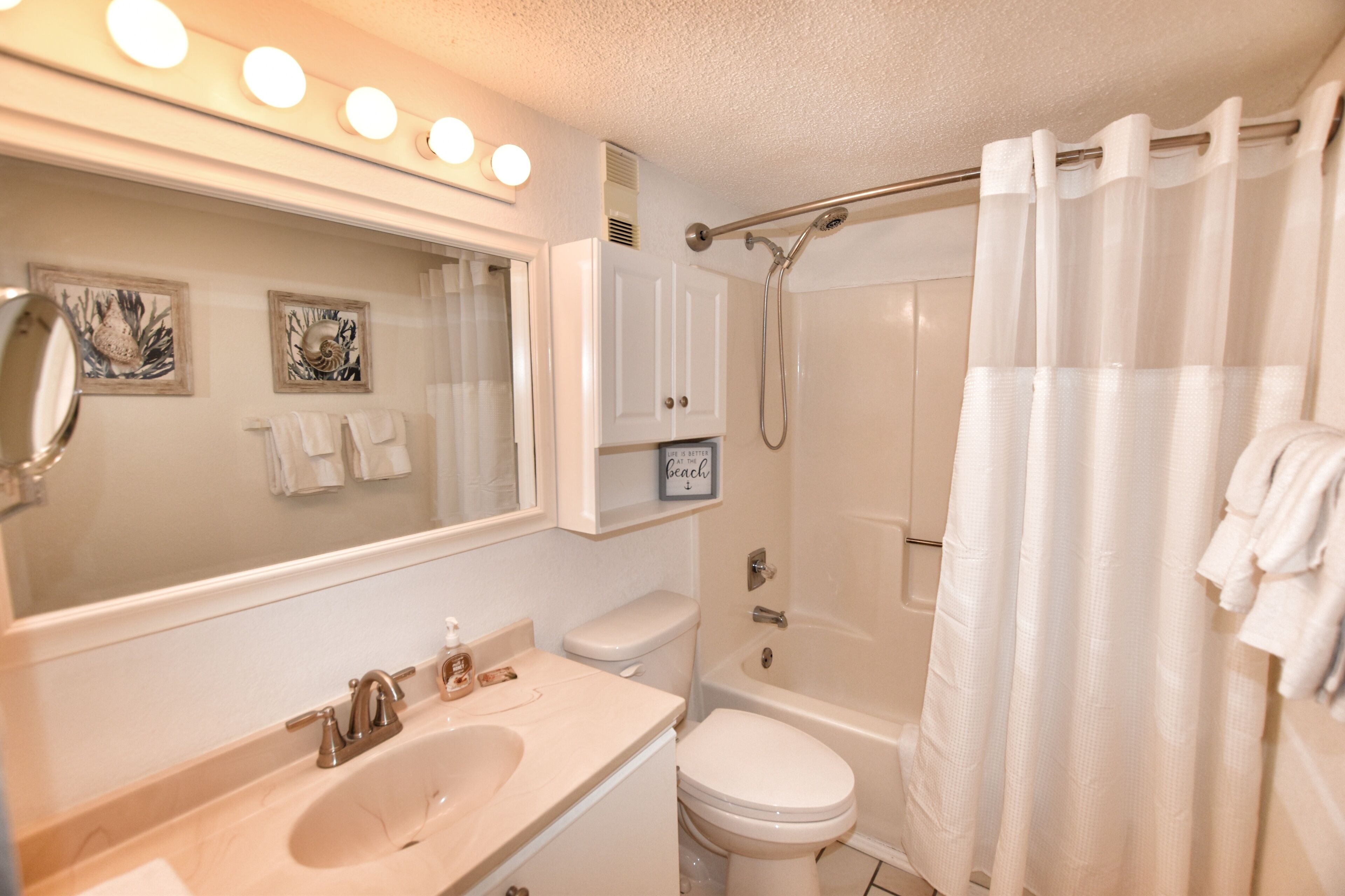 Une salle de bain avec un lavabo, des toilettes et une douche. Il y a des lampes au-dessus du miroir et un porte-serviettes.