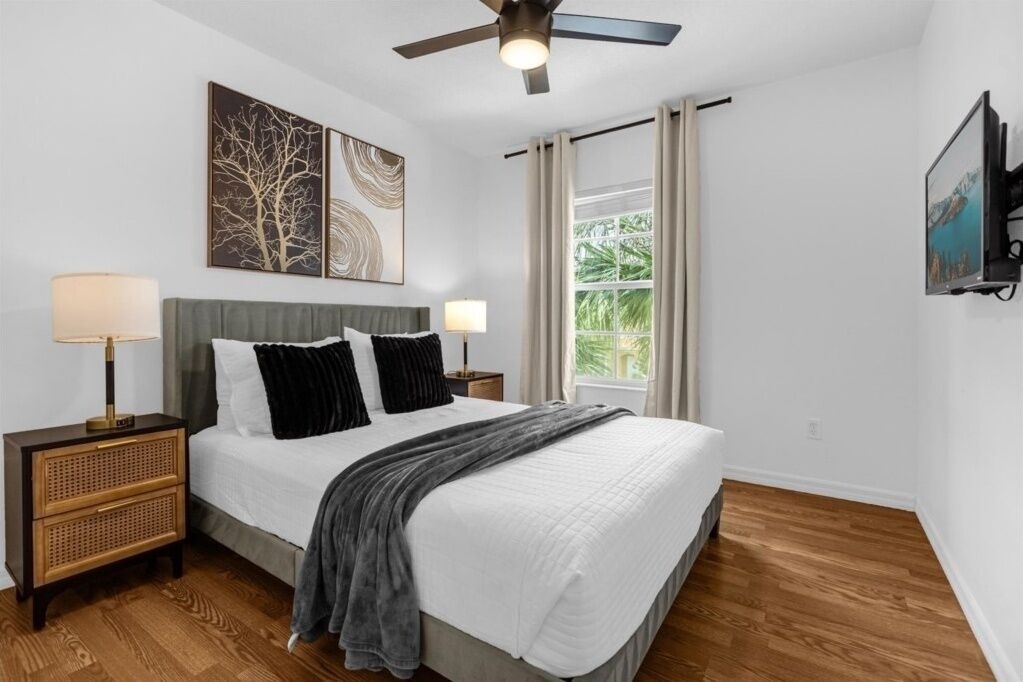 A bedroom with a bed, two bedside tables, a ceiling fan, a window with curtains, and artwork on the wall.