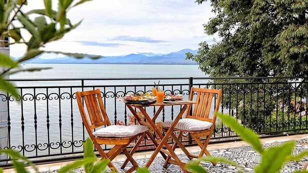 A balcony with a table and two chairs, overlooking a body of water and mountains. - Hotel Labiatan (Shkodër)