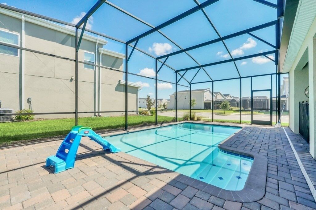 A covered outdoor swimming pool with a blue slide, surrounded by a glass enclosure and brick flooring.