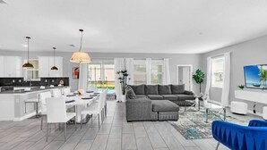 Un salon moderne avec un canapé d’angle gris, un coin repas avec des chaises blanches et une cuisine dotée de meubles blancs et d’un éclairage suspendu.