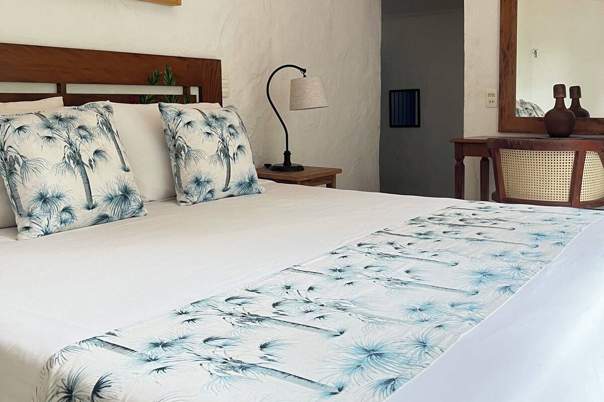 Ein Hotelzimmer mit einem großen Bett, einem Nachttisch mit Lampe, einem Holzstuhl, einem großen Spiegel und einem Gemälde an der Wand.