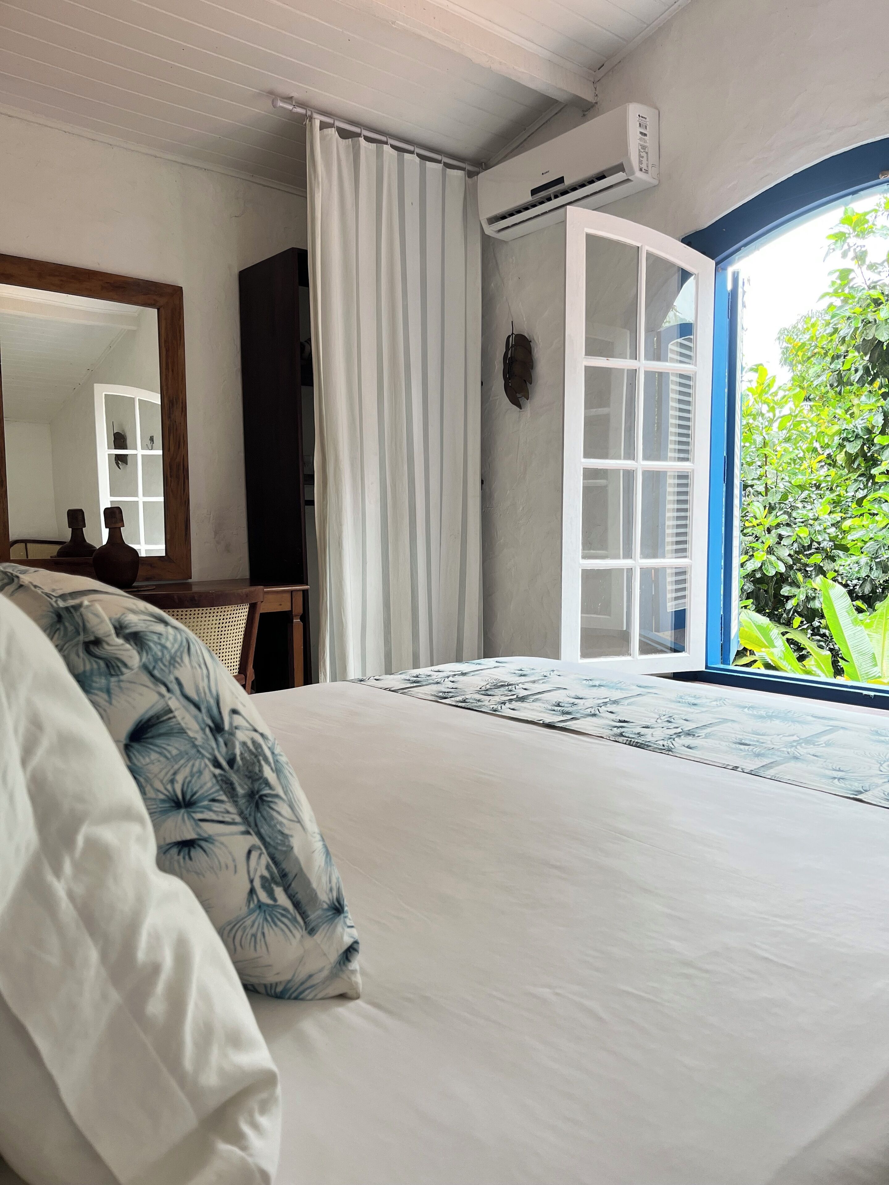 A bedroom with a bed, a mirror, a wardrobe, a window with a view of greenery, and an air conditioning unit.