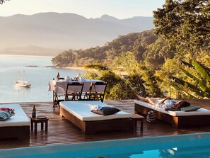 Ein Poolbereich mit Liegestühlen und einem Esstisch, mit Blick auf Strand und Berge.