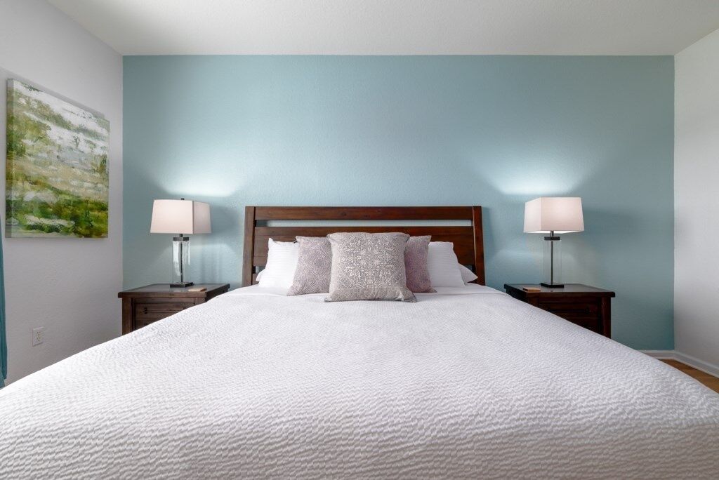 A neatly made bed with a white duvet, two bedside tables with lamps, and a light blue wall with a painting.