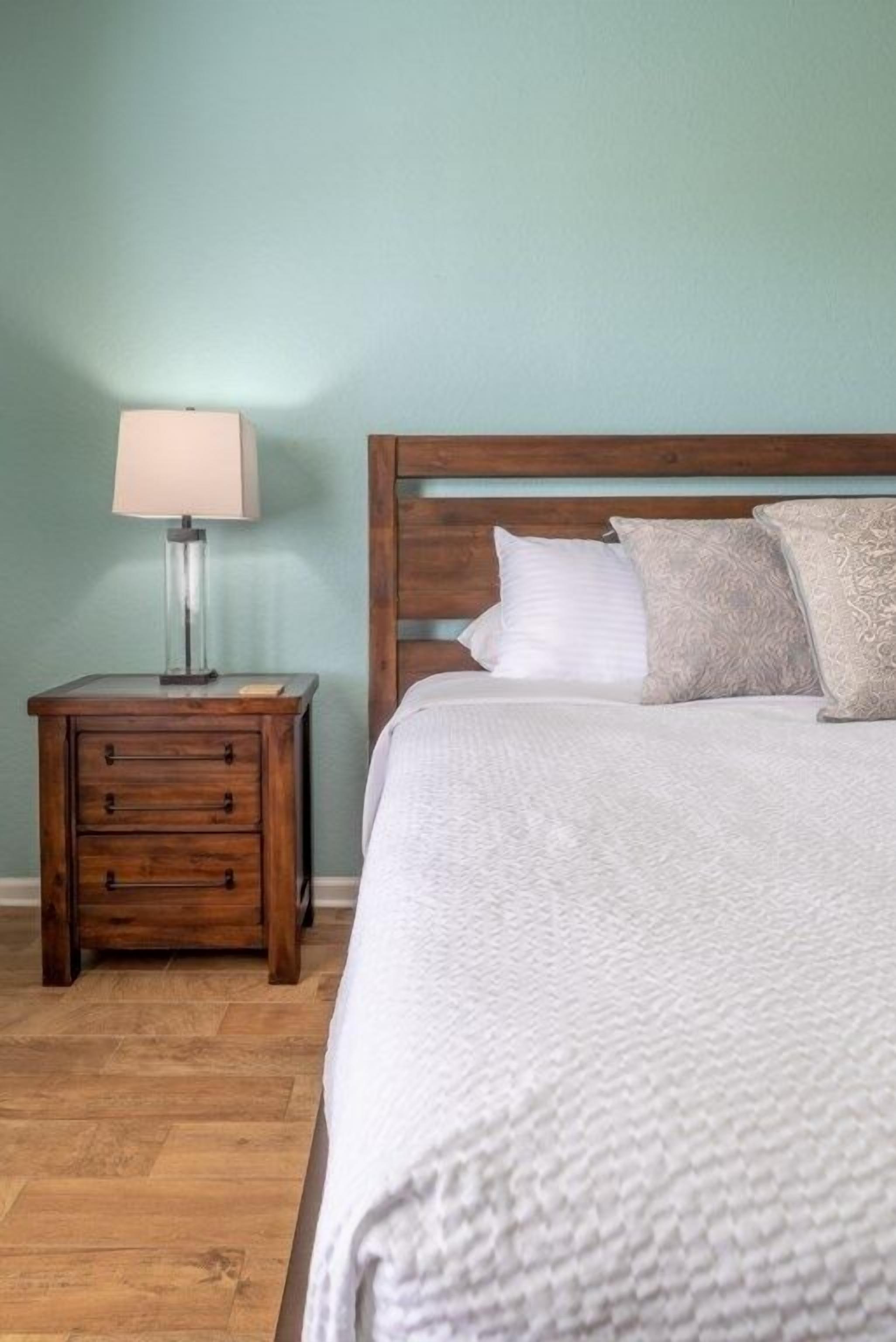 A bedroom with a wooden headboard, a white textured bedspread, a nightstand with a lamp, and a wooden floor.