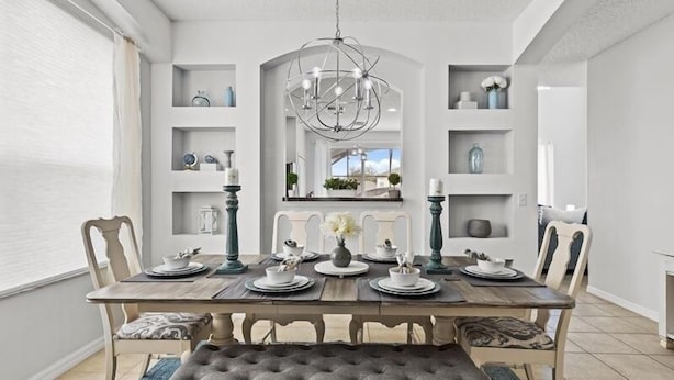 Une salle à manger avec une table en bois dressée pour quatre personnes, entourée de chaises. Un lustre est suspendu au-dessus de la table, et des étagères ornées d’objets décoratifs sont fixées aux murs.