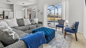 Un salon spacieux avec un grand canapé d’angle gris, un pouf bleu et un fauteuil à motifs. Une grande fenêtre offre une vue sur une piscine et un jardin.