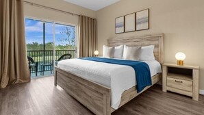 A bedroom with a wooden bed, a nightstand, a balcony with a view of greenery, and two framed pictures on the wall. - Oakwater Retreat 3 Bedroom Condo (Kissimmee)