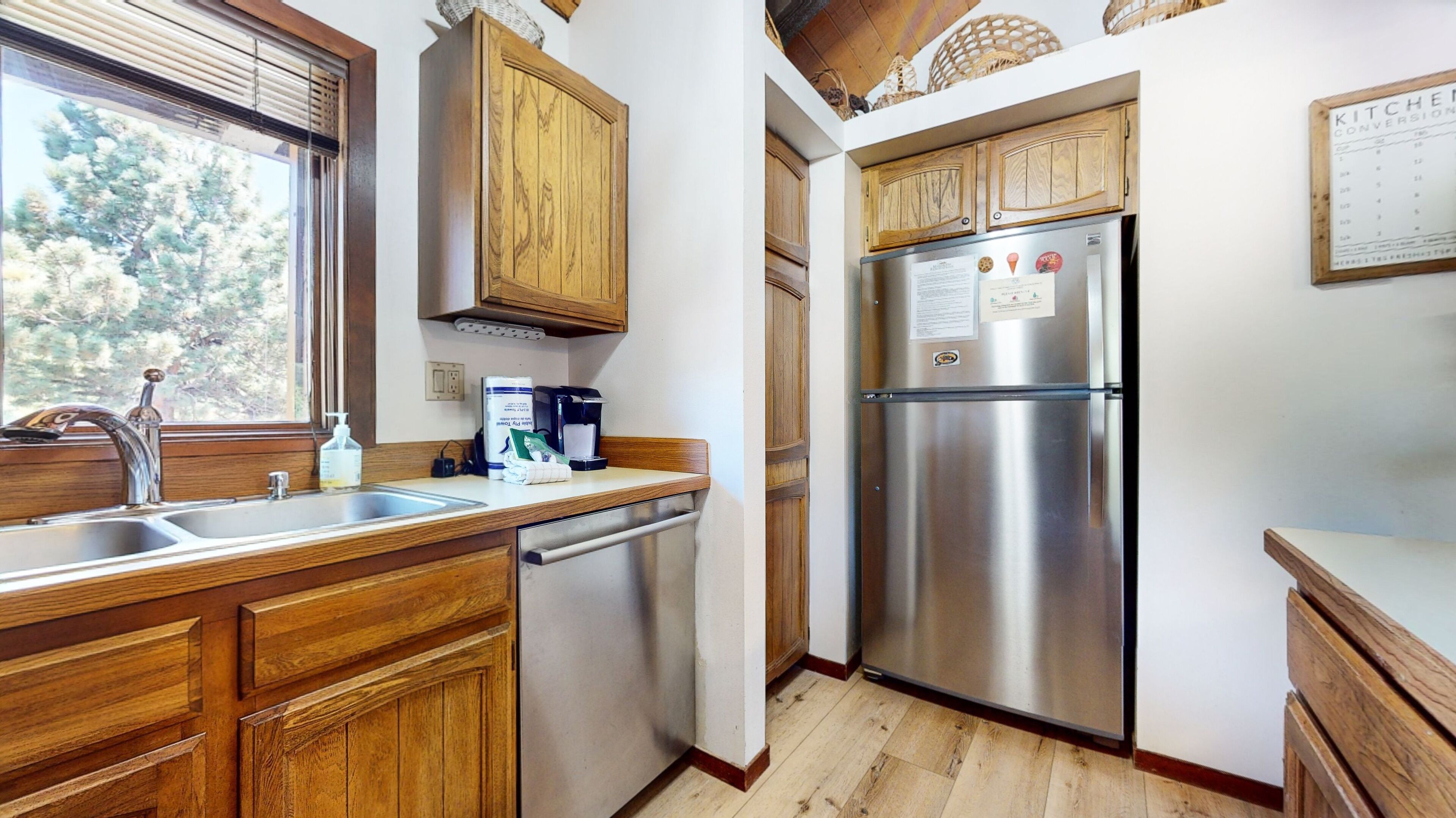 A kitchen with wooden cabinets, a stainless steel refrigerator, a sink, and a window with a view of trees.