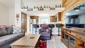 A modern living room with a sofa, a coffee table, and a TV. There is a kitchen area with wooden cabinets and a dining table with chairs. - Forest Creek #16 2 Bedroom Condo by RedAwning (Mammoth Lakes)