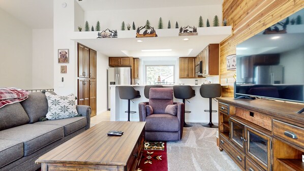 Una sala moderna con sofá, una mesa de centro y televisor. Se observa una cocina con armarios de madera y una mesa de comedor con sillas.