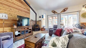 A cozy living room with a TV, fireplace, wooden walls, and a large window. - Forest Creek #16 2 Bedroom Condo by RedAwning (Mammoth Lakes)