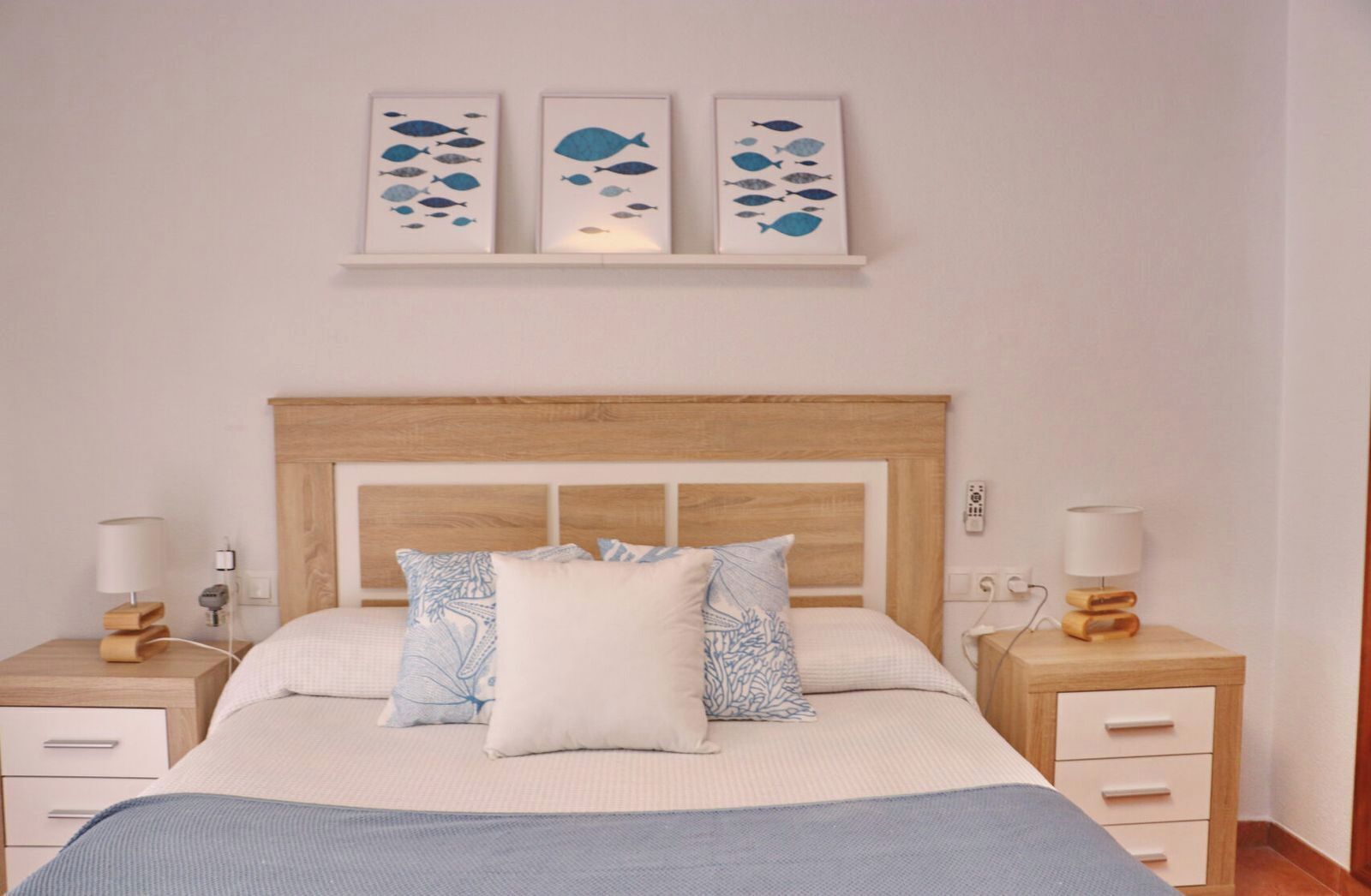 Ein Schlafzimmer mit einem hölzernen Kopfteil, einem Bett mit blau-weißer Bettdecke, zwei Nachttischen mit Lampen und drei an der Wand befestigten Bildern von Fischen.