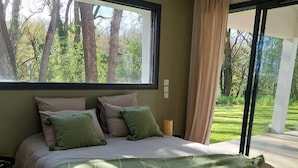 Ein Schlafzimmer mit einem großen Bett, Blick auf Bäume durch ein Fenster und einer Schiebetür, die auf einen Balkon führt.
