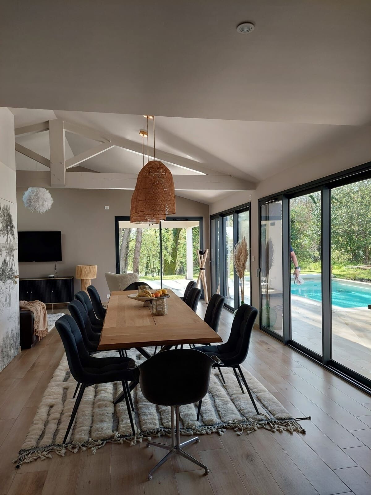 Un comedor moderno con una mesa de madera y sillas negras, ventanas amplias con vista a una piscina y un techo con vigas a la vista.