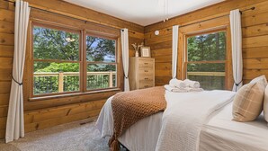 Quarto de cabana de madeira com uma cama, mesa de cabeceira, cômoda e janela com vista para árvores.
