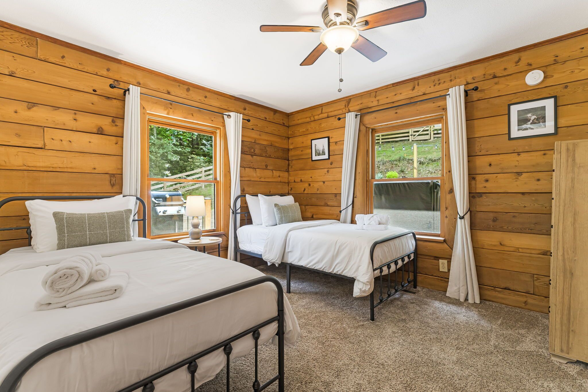 A cozy cabin room with two beds, wooden walls, a ceiling fan, and large windows with curtains.