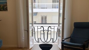 A room with a wooden floor, a dark wooden table with a bowl, a blue chair, and a view of a balcony with white railings. - Newly renovated apartment in Nice- with AC and 10 min walk to the beach! (Nice)