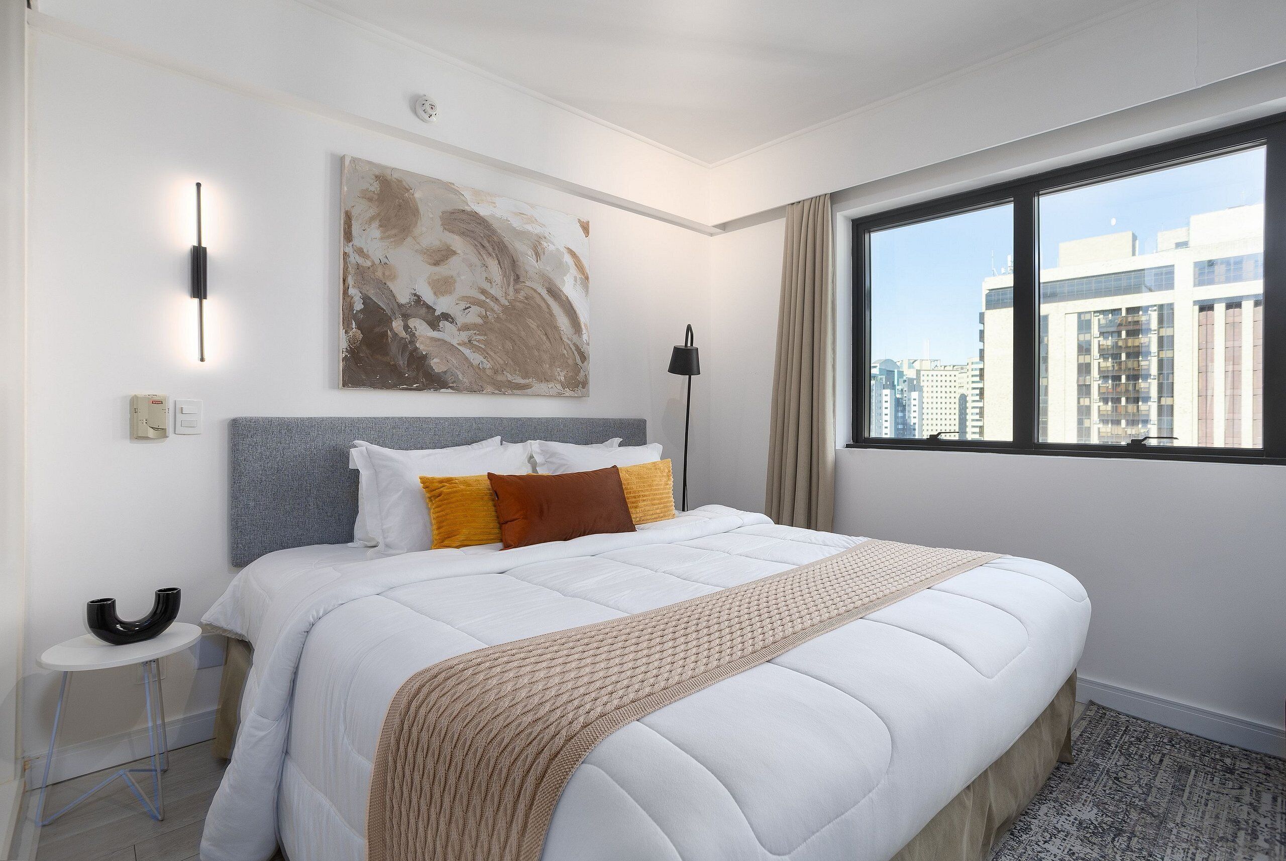 A hotel room with a large bed, a painting on the wall, a window with a city view, and a small table with a lamp.