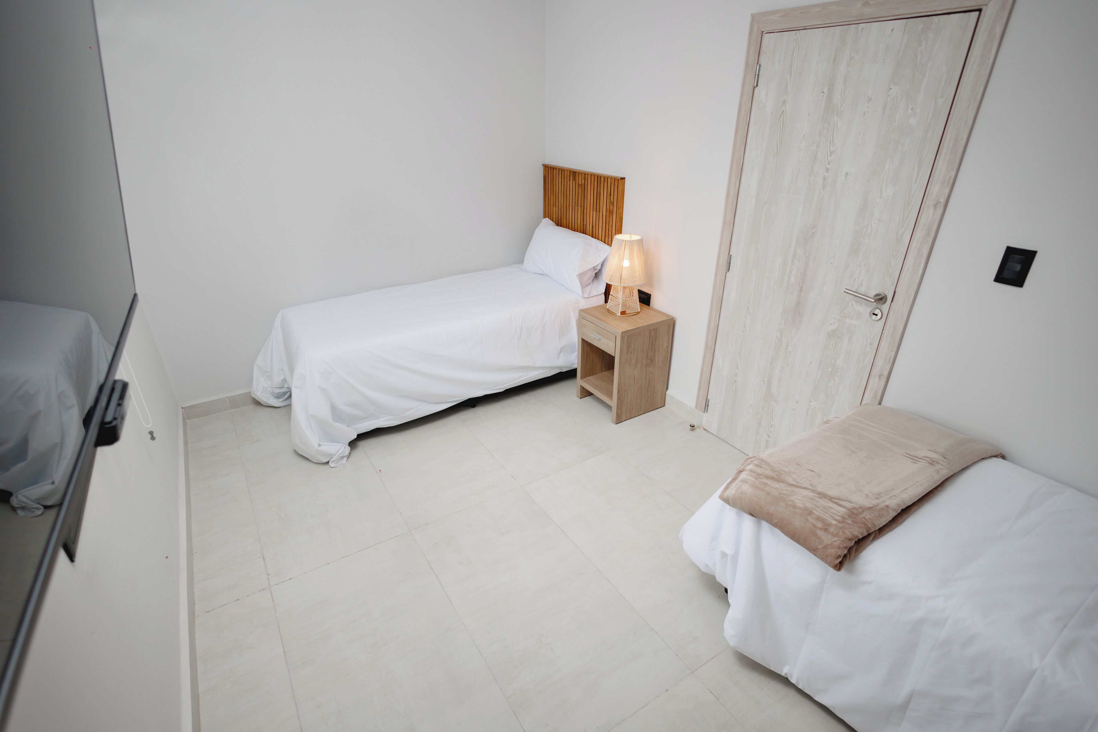 A small, clean room with two single beds, a bedside table with a lamp, and a wooden door.
