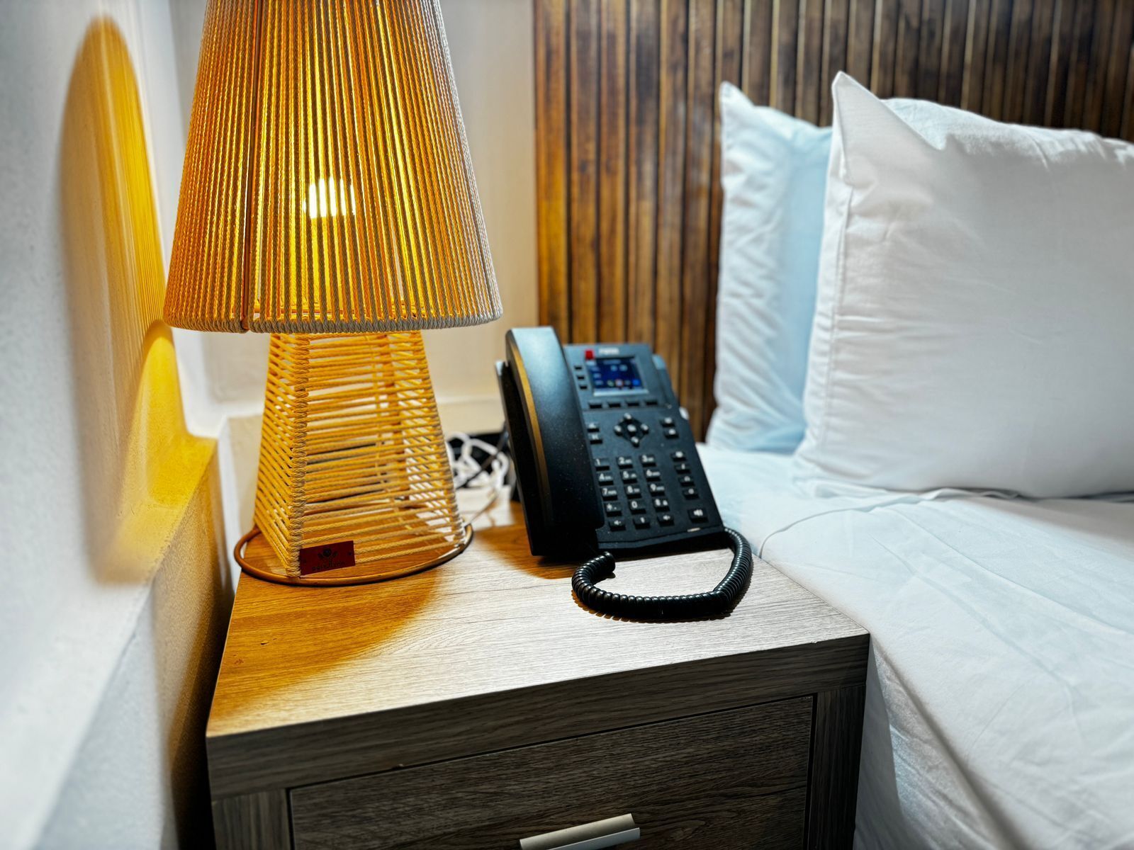 Une table de chevet en bois avec une lampe, un téléphone et un tiroir.