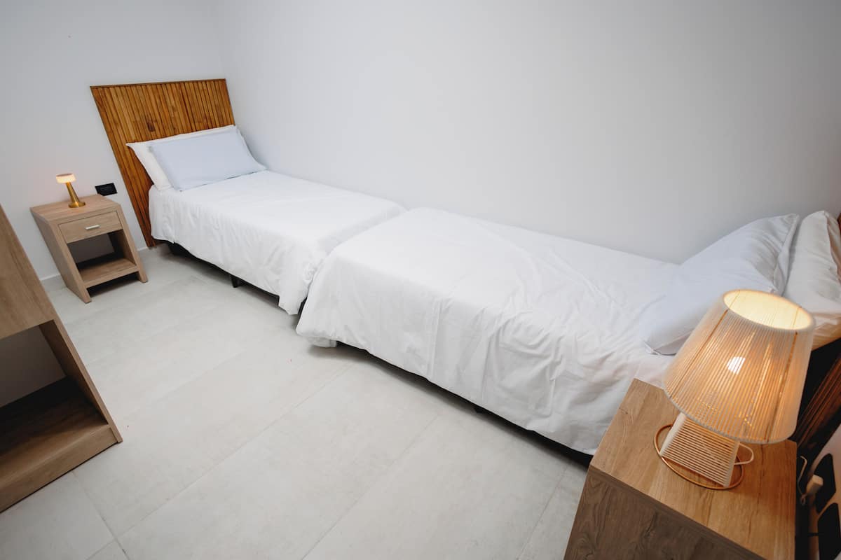 Een enkel bed met wit beddengoed, een houten hoofdbord, een nachtkastje met een lamp, en een houten bijzettafeltje met een lamp.