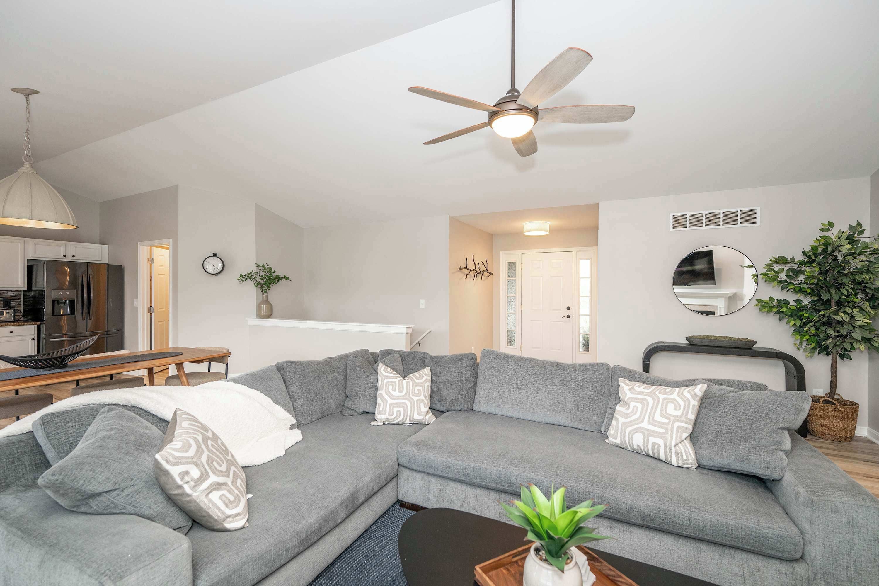 A modern living room with a grey sectional sofa, a ceiling fan, a dining area with a wooden table, and a kitchen in the background.