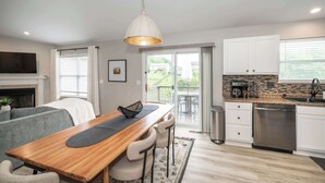 A modern kitchen with a wooden dining table, bar stools, a hanging light fixture, a dishwasher, and a view to an outdoor patio. - Poolside Paradise Near the Ark (Walton)
