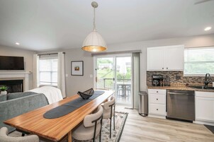 Una cocina moderna con una mesa de comedor de madera, taburetes de bar, una lámpara colgante, lavavajillas y vistas a un patio exterior.