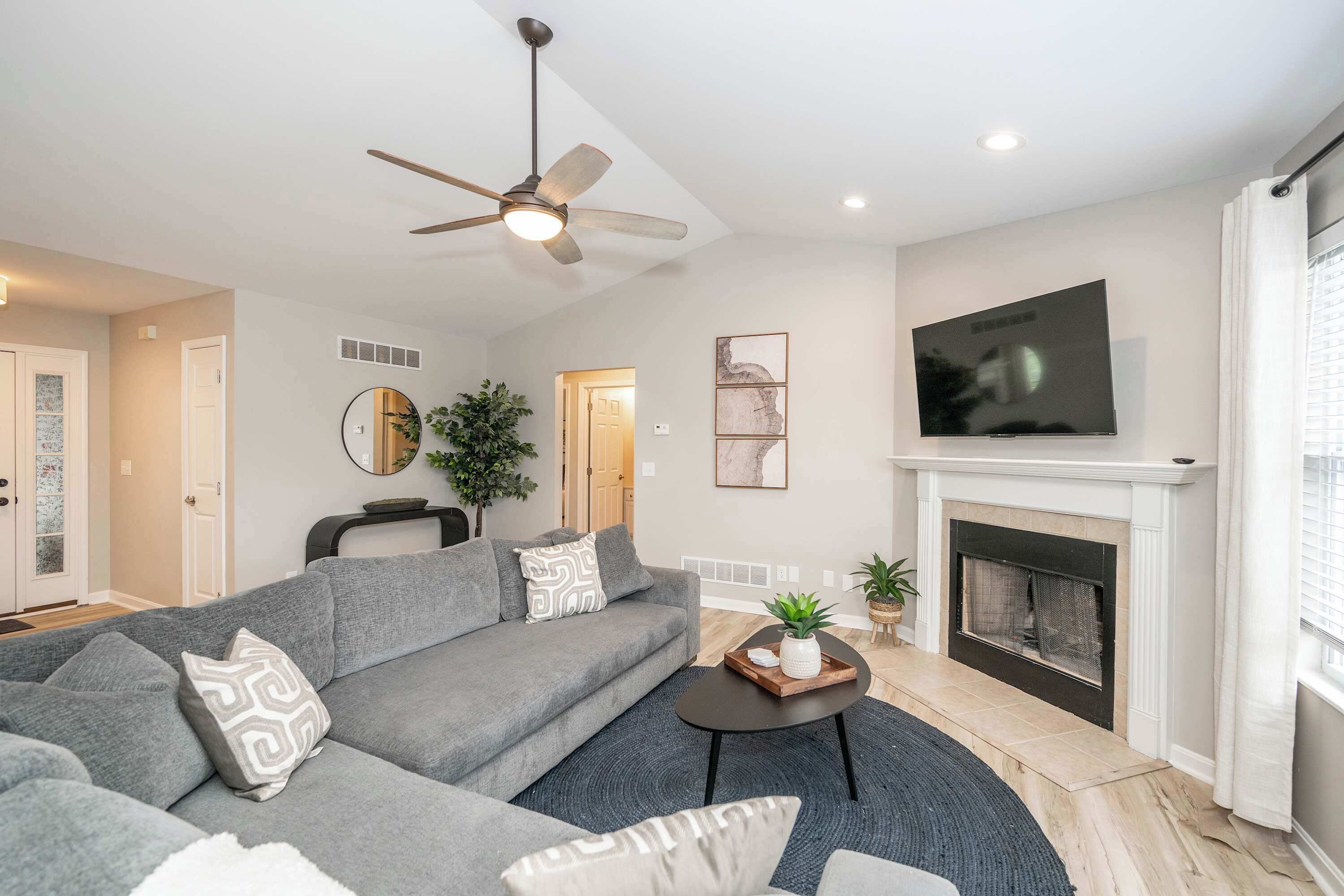 A modern living room with a grey sofa, a fireplace, a ceiling fan, and a flat-screen TV.