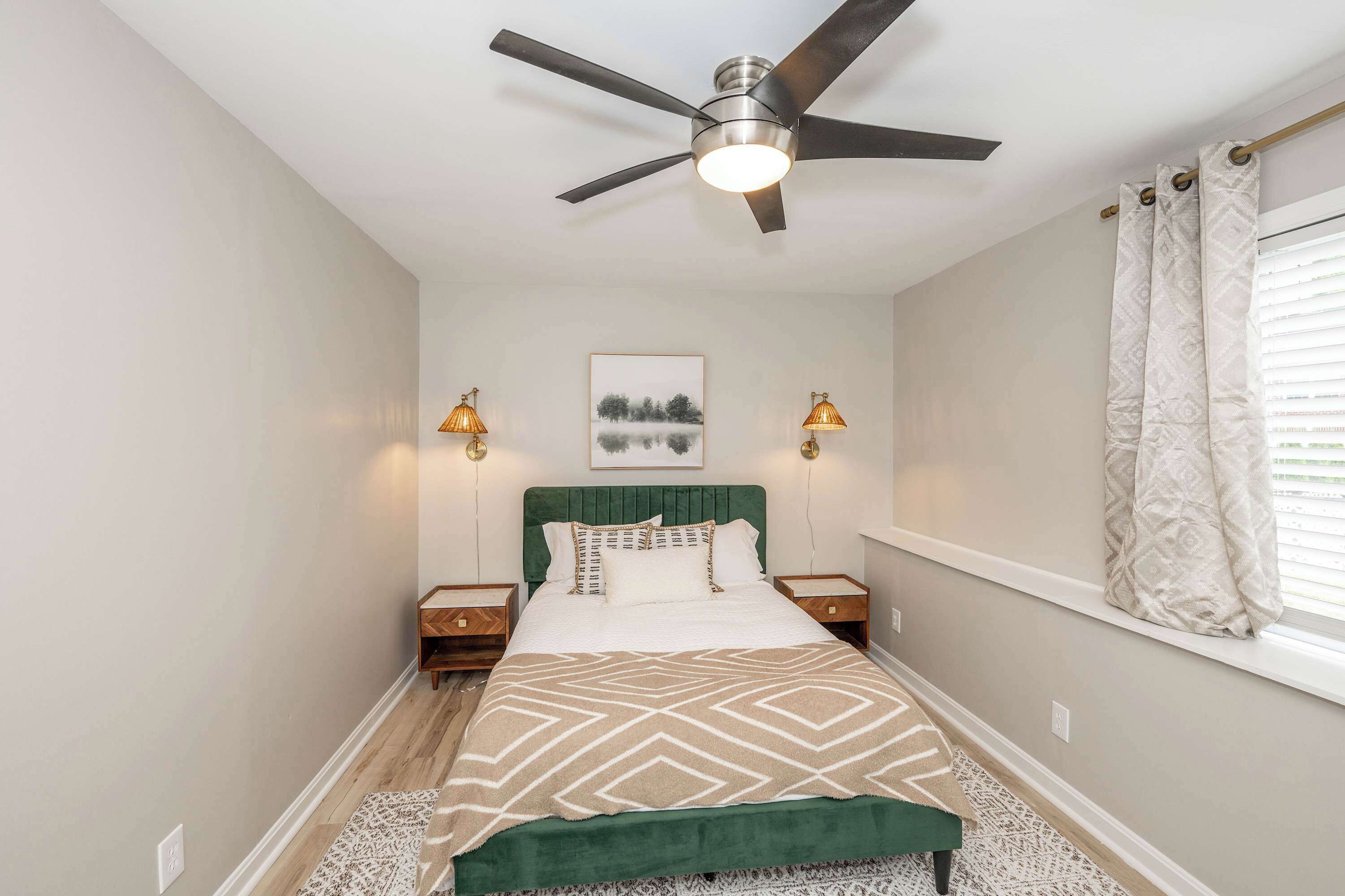 Une chambre à coucher comprenant un lit, un ventilateur de plafond, des lampes de chevet, une fenêtre avec des rideaux et un tableau accroché au mur.