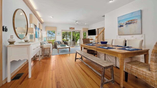 A spacious living area with a wooden floor, a dining table set for four, a large mirror, and a painting of boats on the wall.