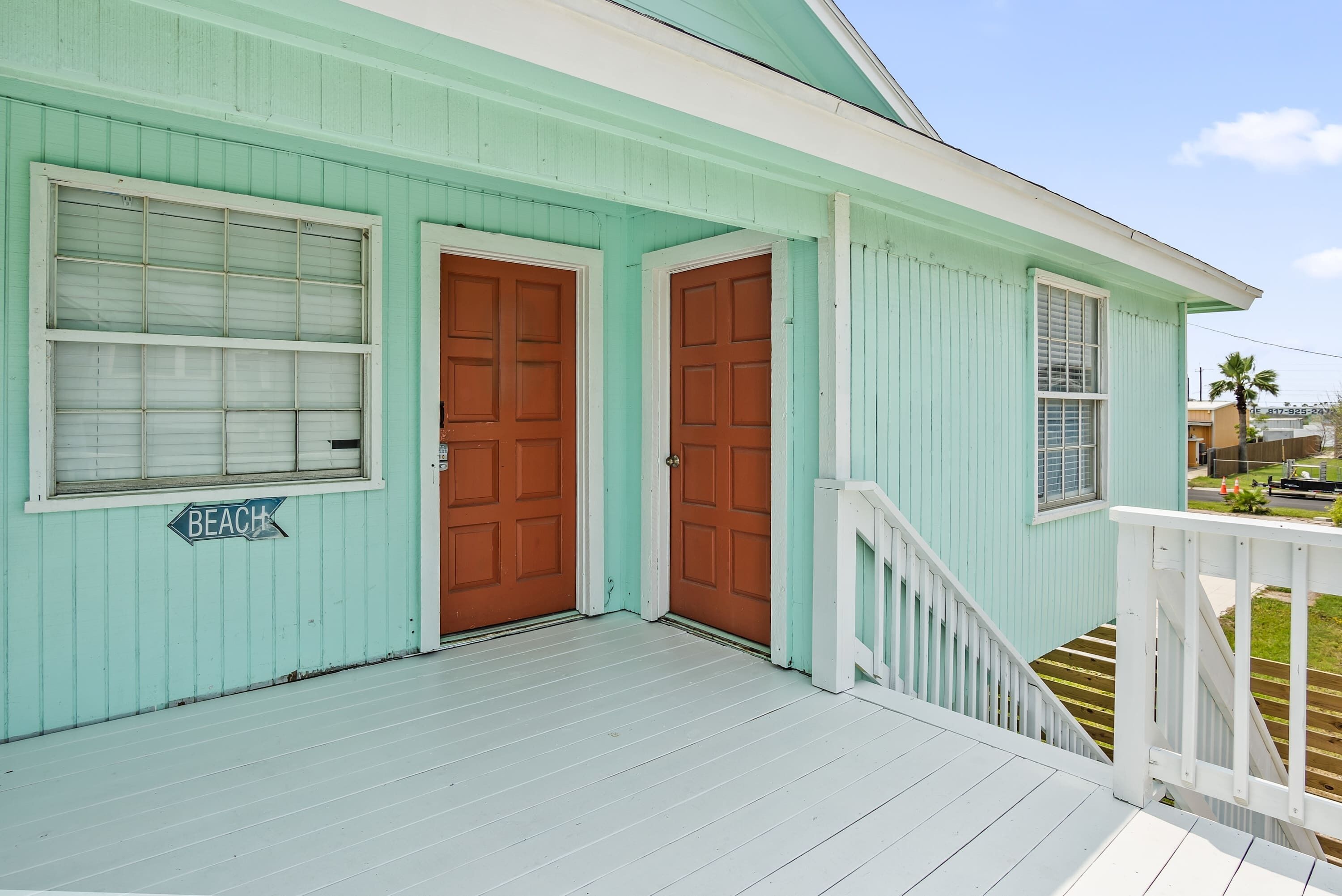 Uma casa azul clara com varanda, duas portas marrons e um letreiro com a palavra 'BEACH'.