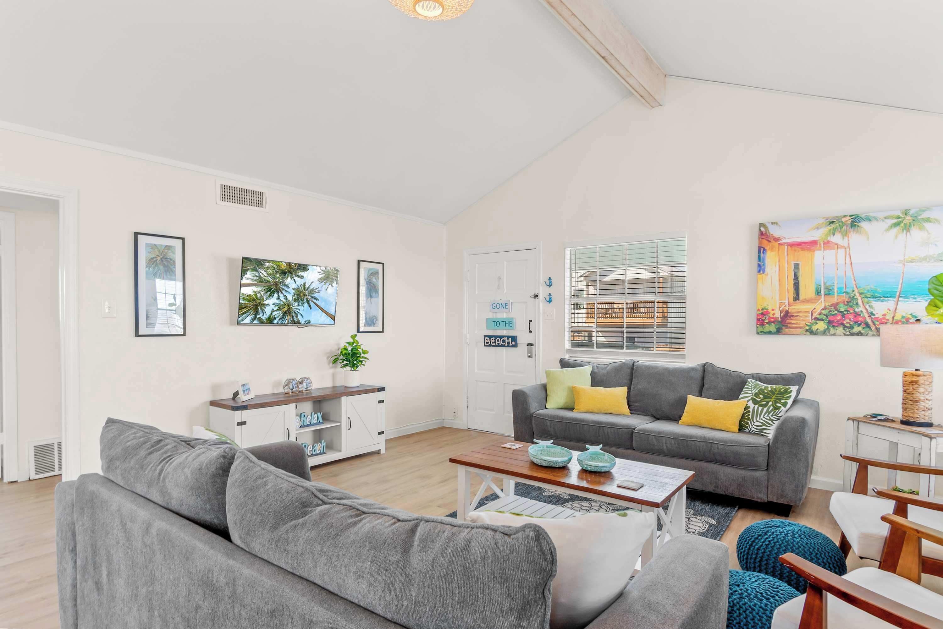 A spacious living room with a grey sofa, a coffee table, and a bookshelf. There is a painting of a beach scene on the wall.