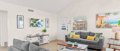 A spacious living room with a grey sofa, a coffee table, and a bookshelf. There is a painting of a beach scene on the wall.