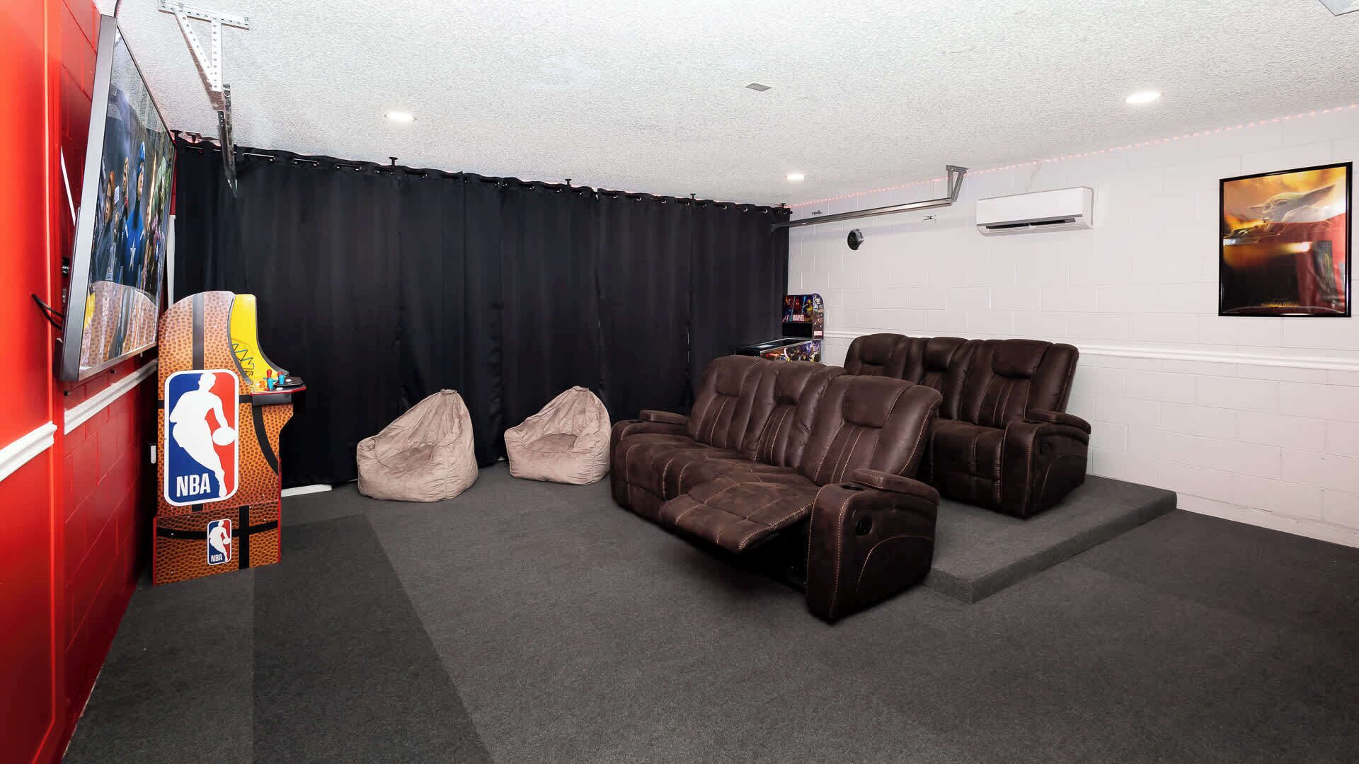 Une salle de cinéma maison avec un mur rouge, des rideaux noirs et un grand écran. On y trouve deux fauteuils inclinables et deux poufs.