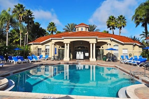 A pool area with a building that has a red-tiled roof and columns, surrounded by palm trees and blue lounge chairs.