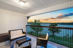 A balcony with wicker furniture, a view of a lake at sunset, and a railing.