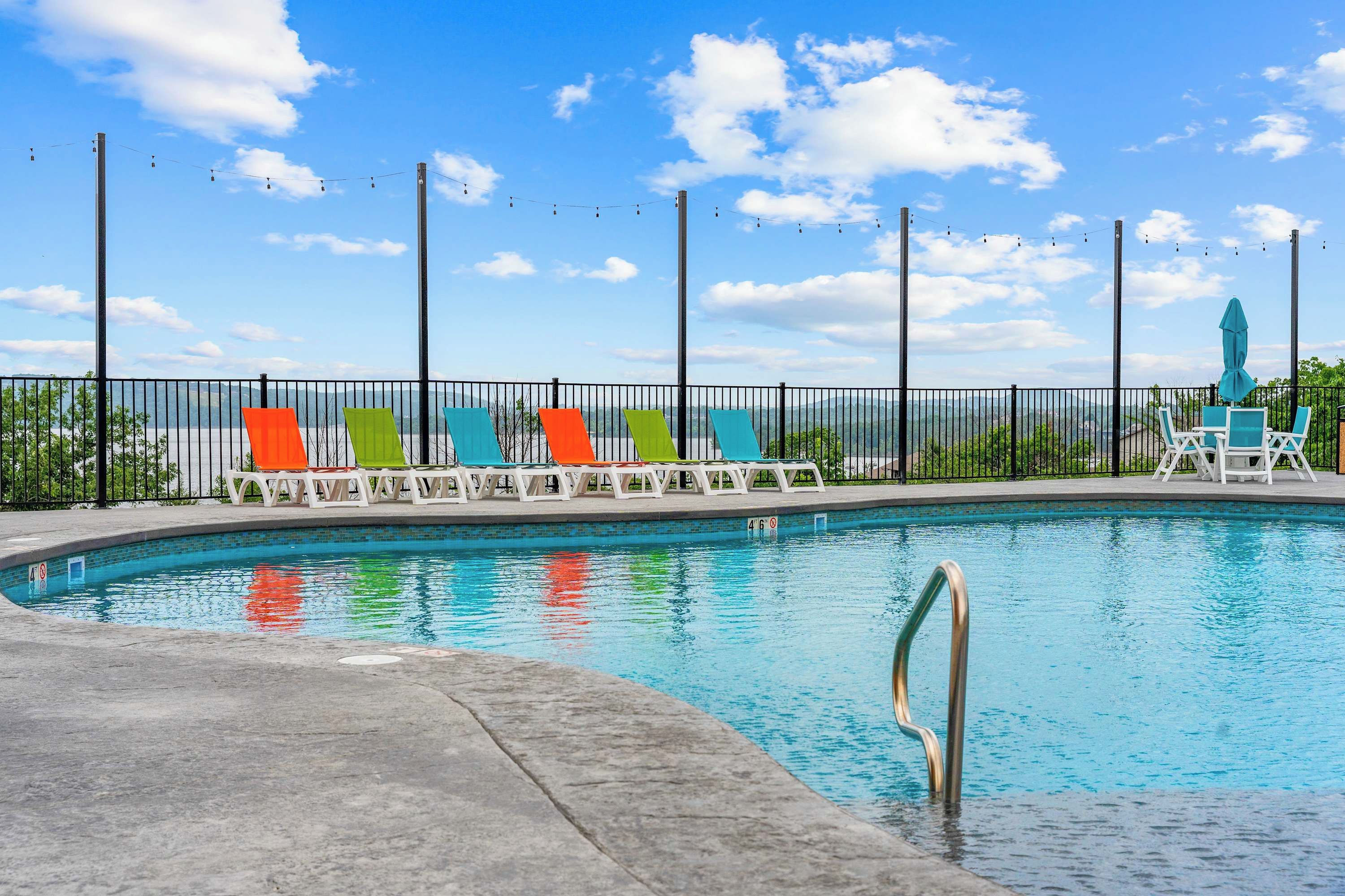 Una piscina con una escalera metálica, rodeada por una cerca y sillas de descanso coloridas.