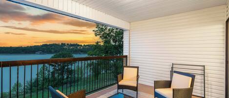 A balcony with wicker furniture, a glass table, and a view of a lake and trees at sunset.