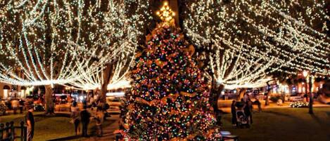 Un gran árbol de Navidad adornado con luces y adornos, rodeado de cajas de regalo, en un espacio público al aire libre.