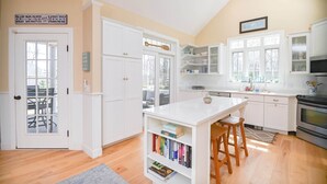 Una cocina luminosa con armarios blancos, una isla central con taburetes y vistas al exterior a través de amplias ventanas.