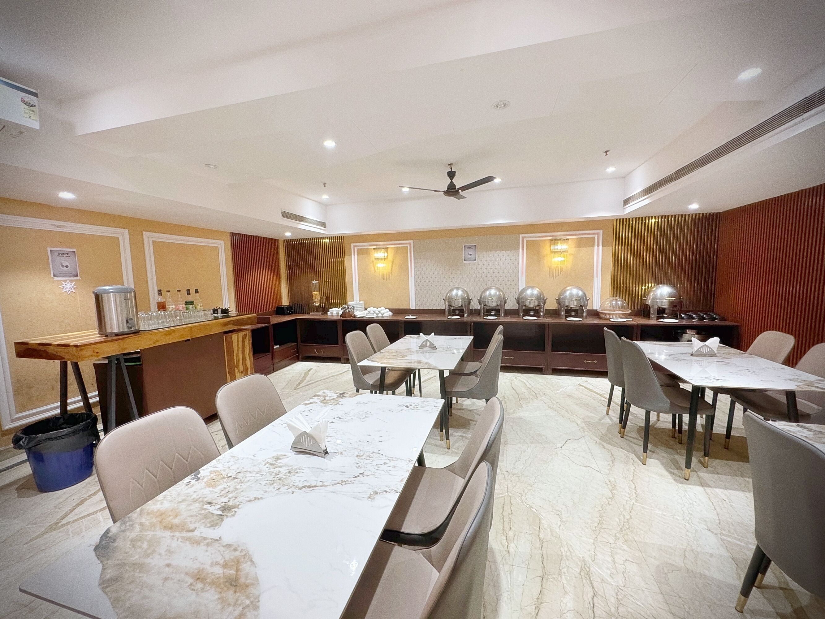Un espace de restauration d’hôtel avec des tables surmontées de marbre, des chaises rembourrées, un buffet et un ventilateur de plafond.