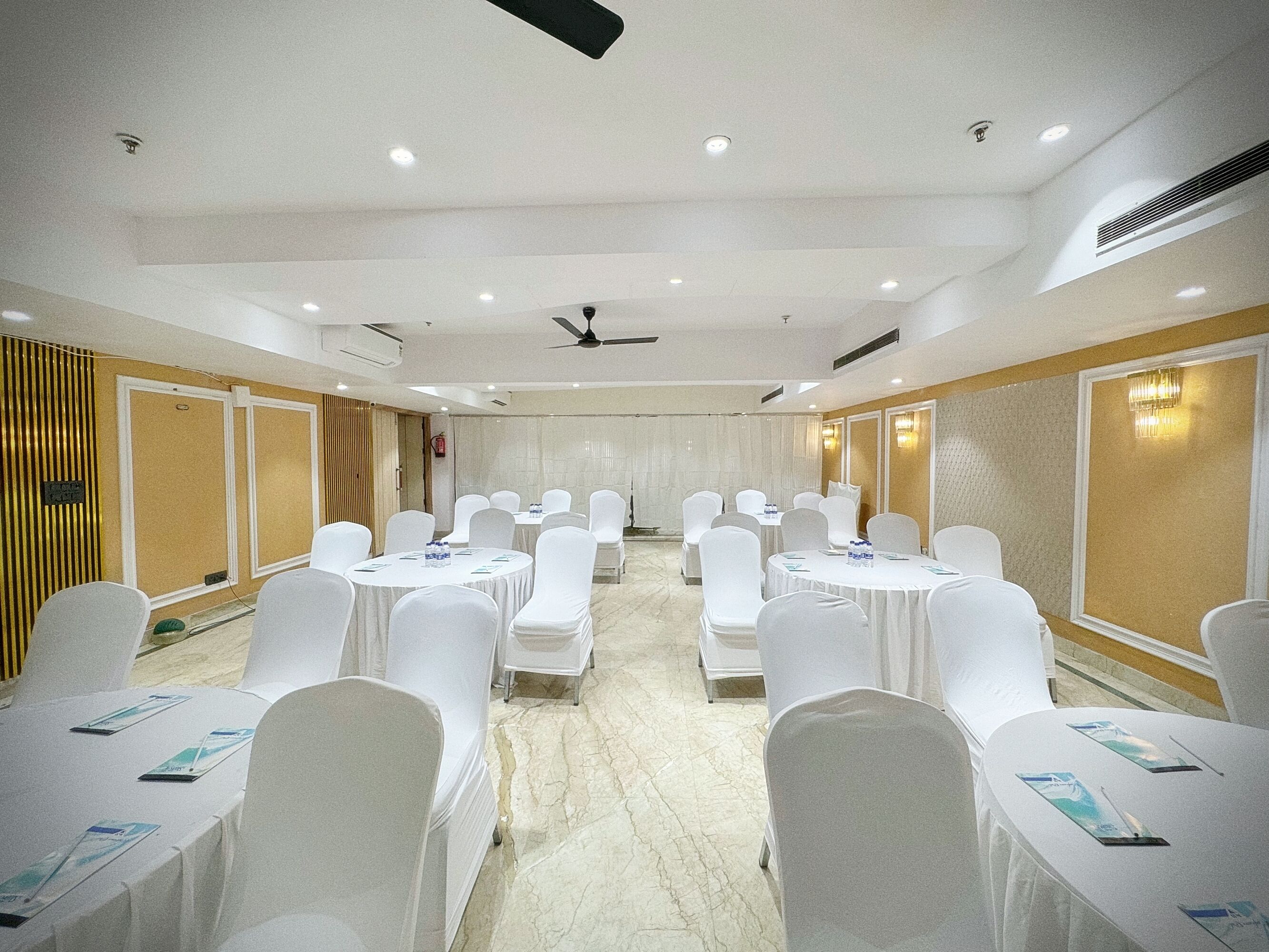 Une salle de conférence dotée de tables rondes et de chaises blanches, équipée de ventilateurs de plafond et d’un système d’éclairage encastré.