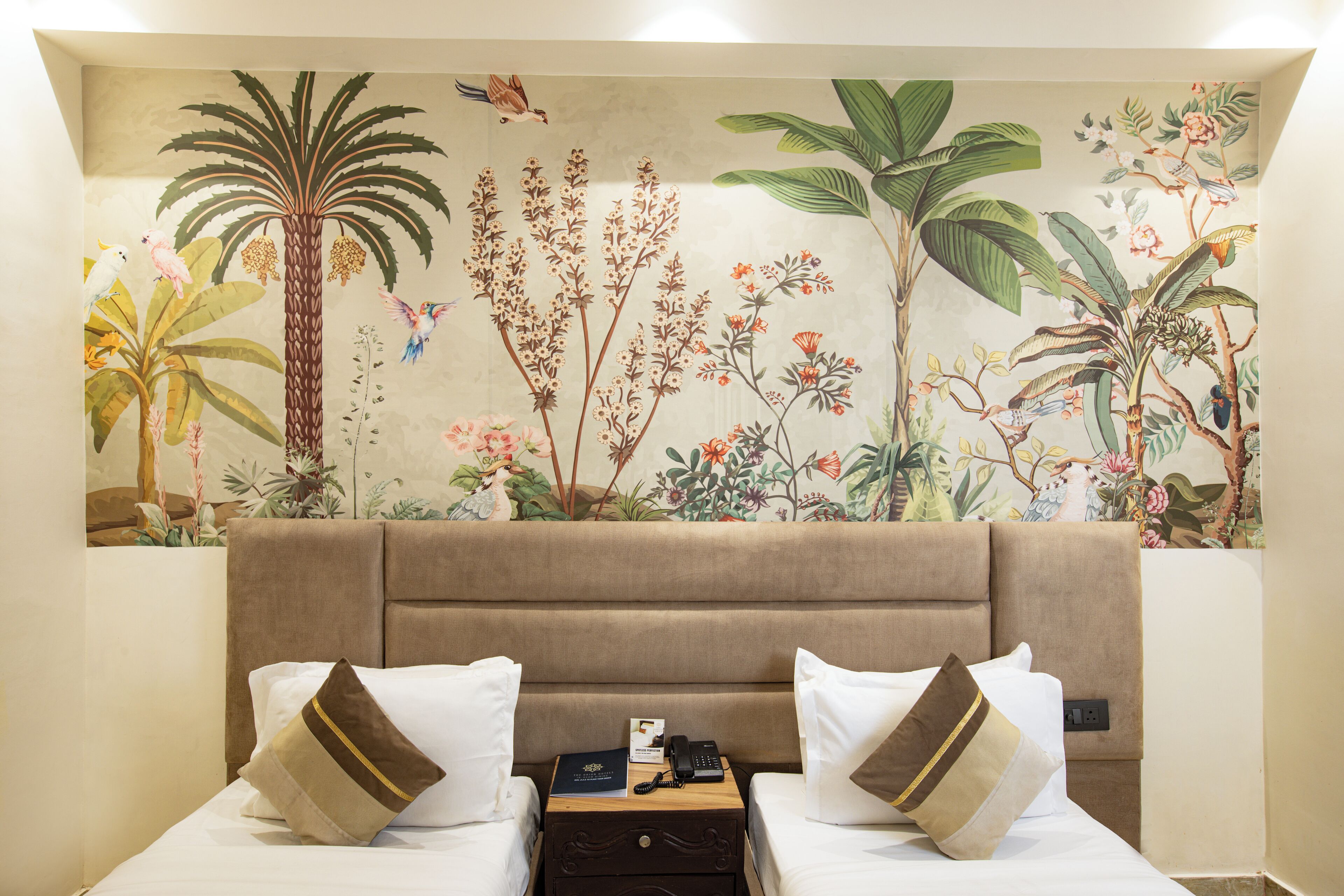 A hotel room with a large floral mural on the wall, two beds with white linens and brown pillows, a nightstand with a phone and a book, and a brown upholstered headboard.
