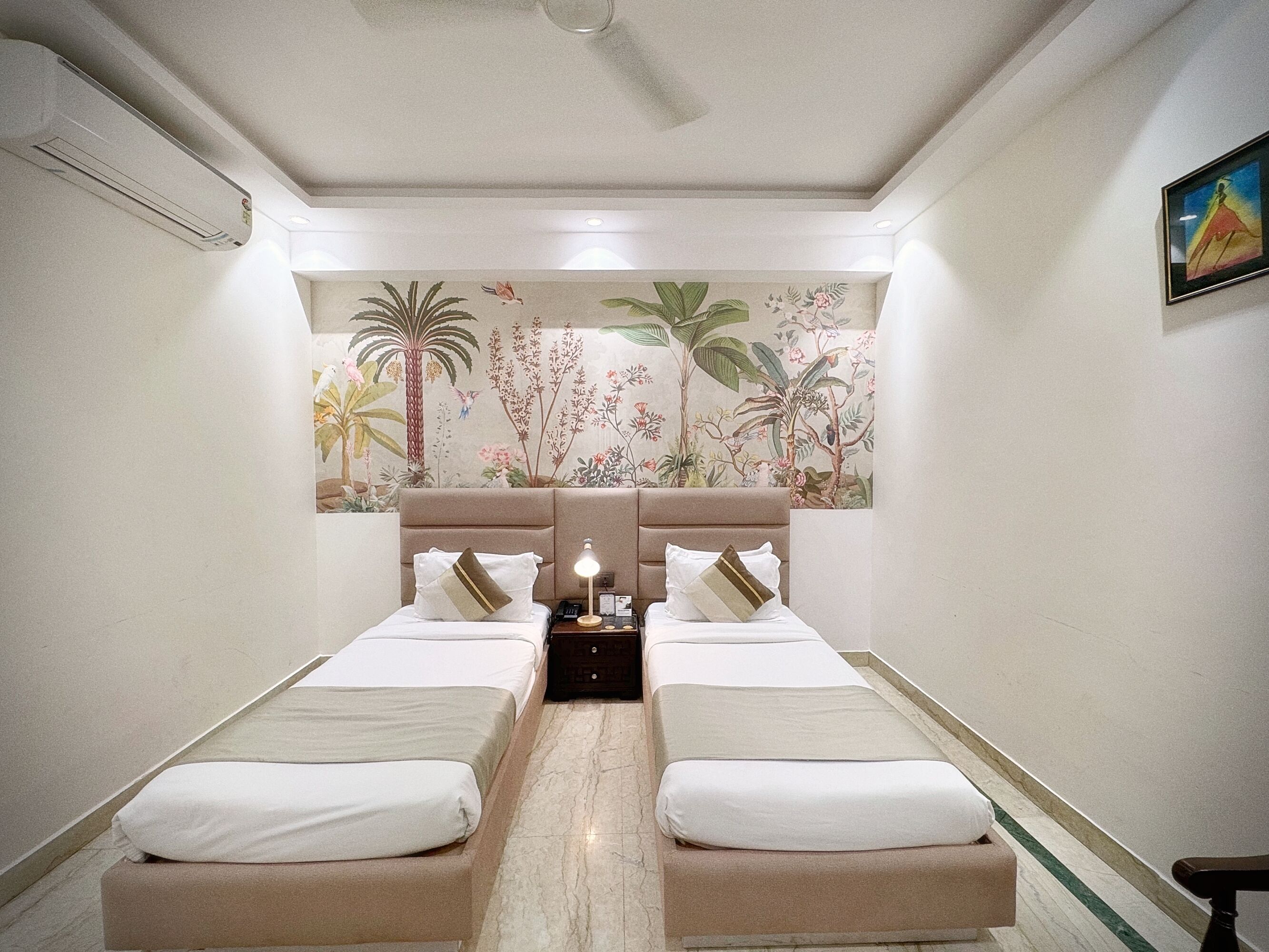 A hotel room with two beds, a ceiling fan, a nightstand with a lamp, and a floral mural on the wall.