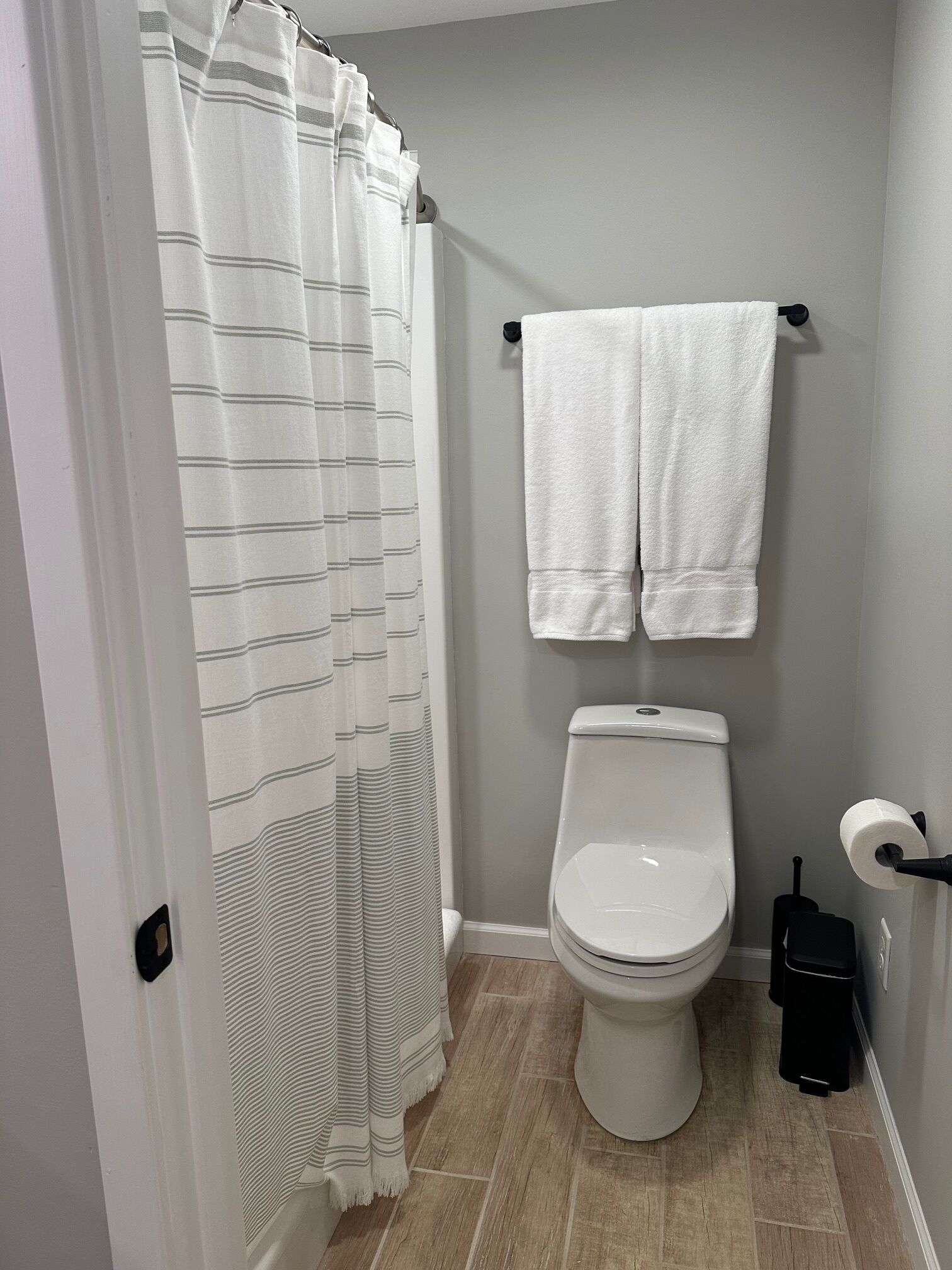 Une salle de bain comprenant des toilettes, un porte-serviettes avec deux serviettes, un rideau de douche à rayures et une poubelle noire.